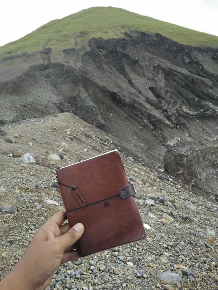 Tangan memegang jurnal kulit Hibrkraft di lereng gunung berbatu.